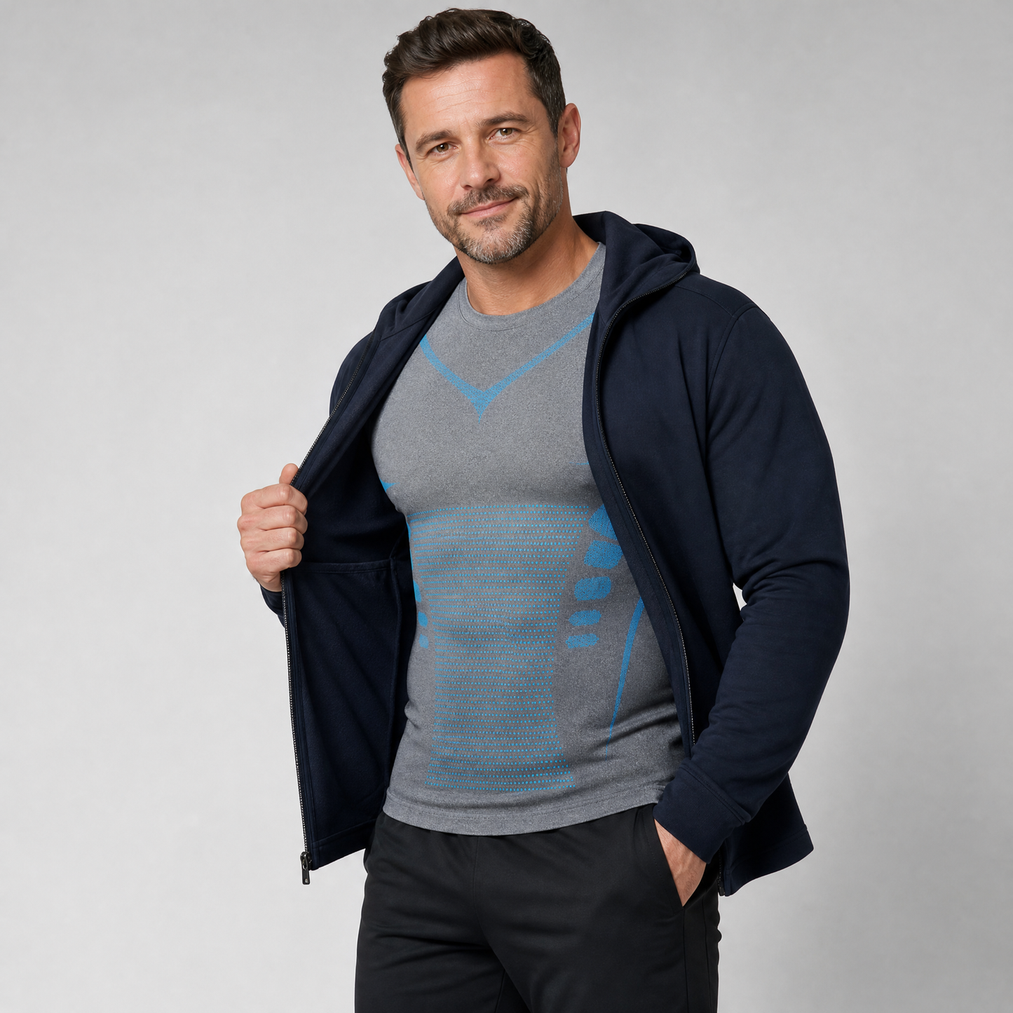 A man wearing a navy blue zip-up hoodie over a Formyx tank top, paired with black pants, standing against a light gray studio background.